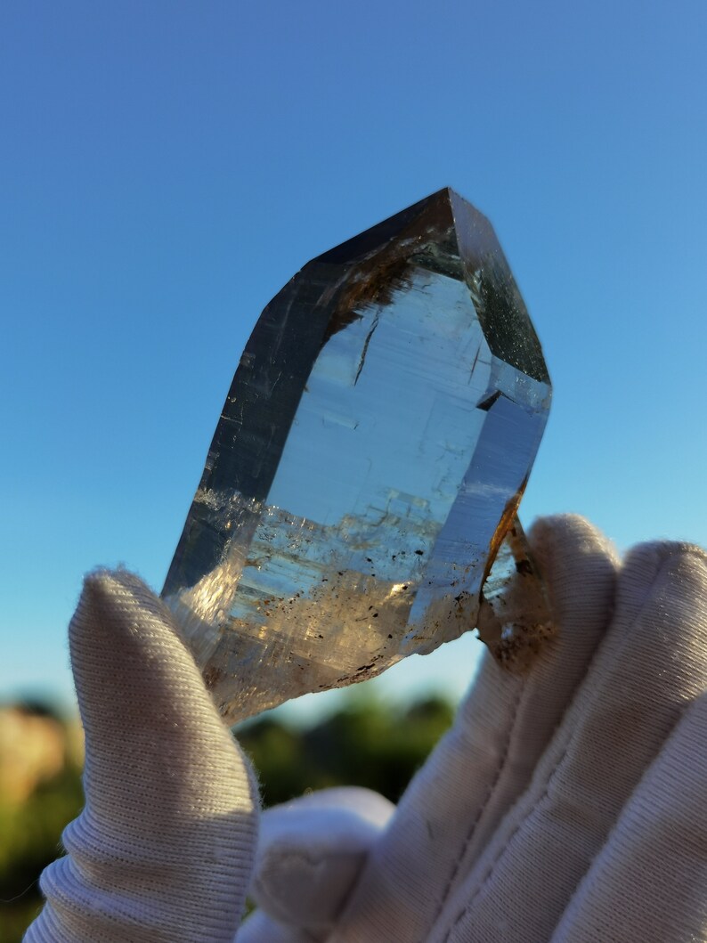 May include: A large, clear quartz crystal with a point, held in gloved hands. The crystal has a slight brown tint and is translucent.