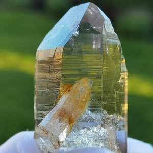 May include: A clear quartz crystal with a golden yellow inclusion. The crystal is pointed at the top and has a flat base. It is being held in a white cloth.