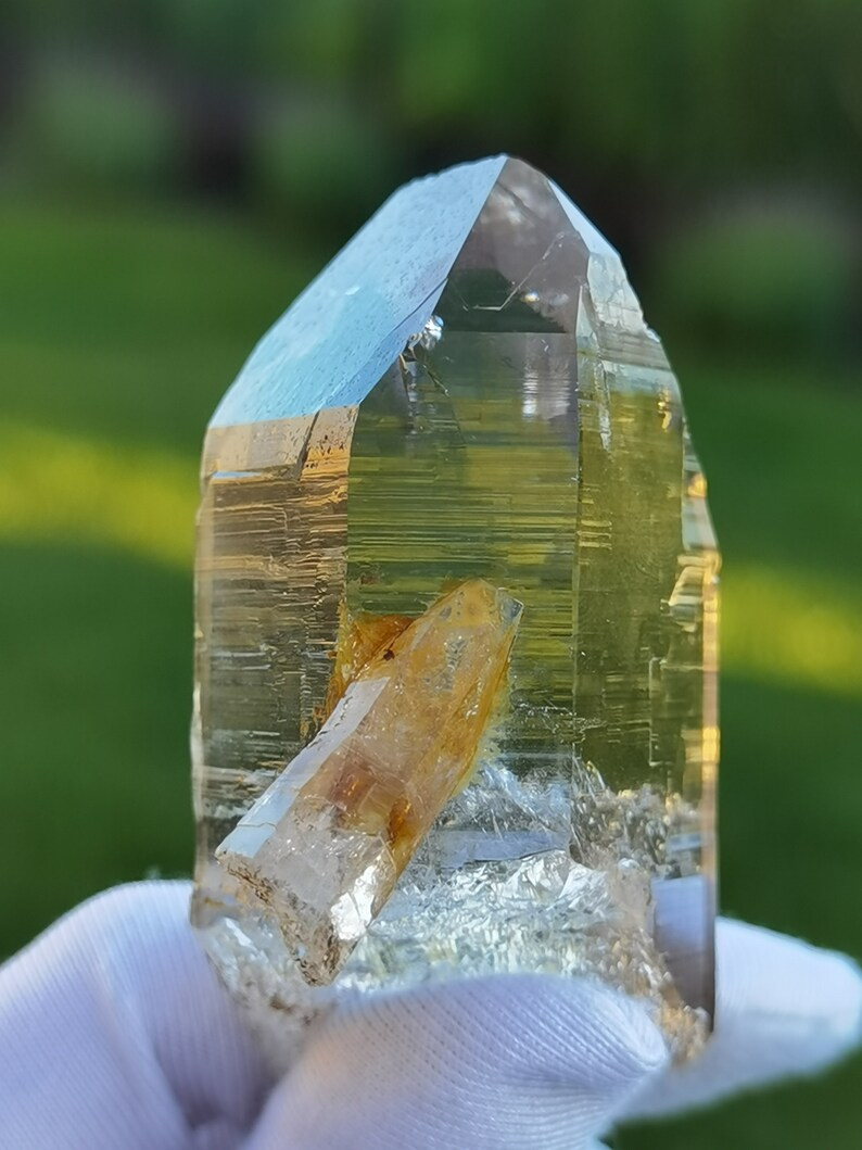 May include: A clear quartz crystal with a golden yellow inclusion. The crystal is pointed at the top and has a flat base. It is being held in a white cloth.