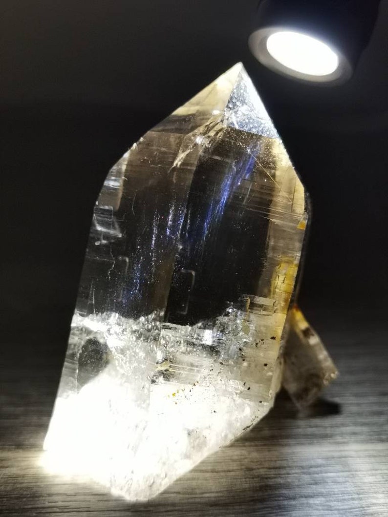 May include: A large, clear quartz crystal with a pointed tip and a flat base. The crystal is illuminated by a light source, highlighting its internal structure and facets.