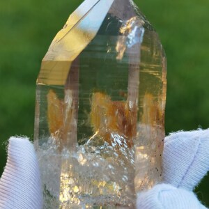 May include: A clear quartz crystal with a golden brown inclusion. The crystal is faceted and has a pointed tip. It is being held in a white gloved hand.