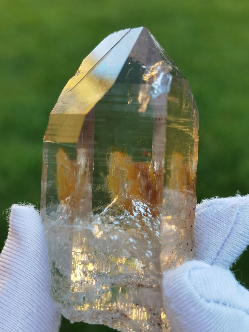 May include: A clear quartz crystal with a golden brown inclusion. The crystal is faceted and has a pointed tip. It is being held in a white gloved hand.