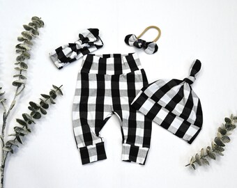May include: A set of baby clothing featuring a black and white buffalo plaid pattern. The set includes a pair of cuffed leggings, a knotted beanie hat, a wide fabric headband with a bow, and a thin elastic headband with a small bow.