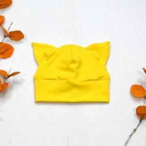 May include: A yellow knitted hat with cat ears. The hat is made of a soft, stretchy fabric.
