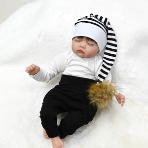 May include: A sleeping baby doll wearing a white long-sleeved shirt, black pants, and a white and black striped hat with a brown pom-pom. The doll is lying on a white fluffy surface.
