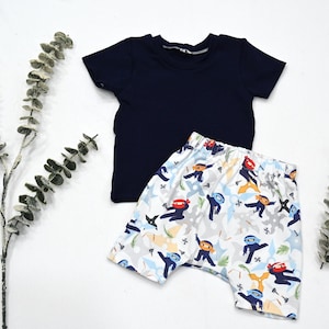 May include: A navy blue short-sleeved t-shirt and a pair of patterned shorts. The shorts feature a ninja print in red, blue, and yellow on a white background. The clothing set is likely for a child.