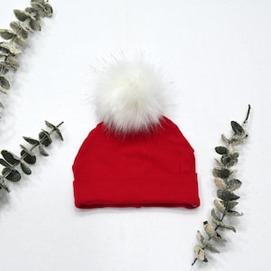 May include: Red knit beanie with a white faux fur pom pom. The beanie is on a white background.