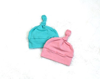May include: Two soft fabric baby hats, one in a solid light turquoise color and the other in a solid light pink color. Each hat features a folded brim at the base and a decorative knot tied at the top center, creating a simple, rounded shape.