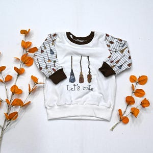 May include: White long-sleeved sweatshirt with brown trim and sleeves patterned with brooms on a striped background. The front features three broom illustrations and the text "Let's ride".