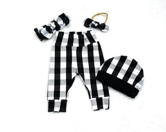 May include: A set consisting of black and white buffalo plaid trousers with a high waistband, a matching beanie with a solid black cuff, a wide fabric headband with a bow, and a smaller bow headband on a thin tan elastic band.
