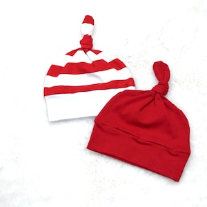 May include: Two red and white striped and solid red knotted baby hats. The hats are made of soft fabric and are designed to fit newborns. The hats are on a white background.