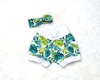 May include: A pair of shorts featuring a white background with a green and dark green tropical leaf print, finished with solid white fabric at the waistband and leg cuffs, accompanied by a matching knotted fabric headband.