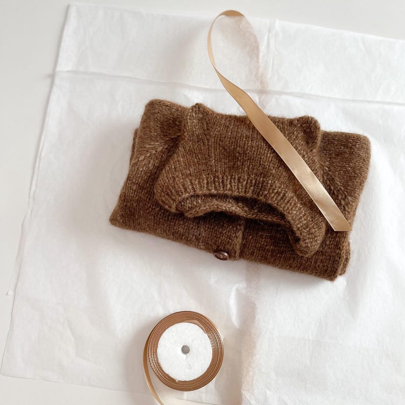 May include: A folded brown knit cardigan with a matching knit hat featuring small rounded ears on top. A single brown button is visible on the cardigan. A spool of light tan ribbon is placed nearby, with a length of the same ribbon draped over the items.