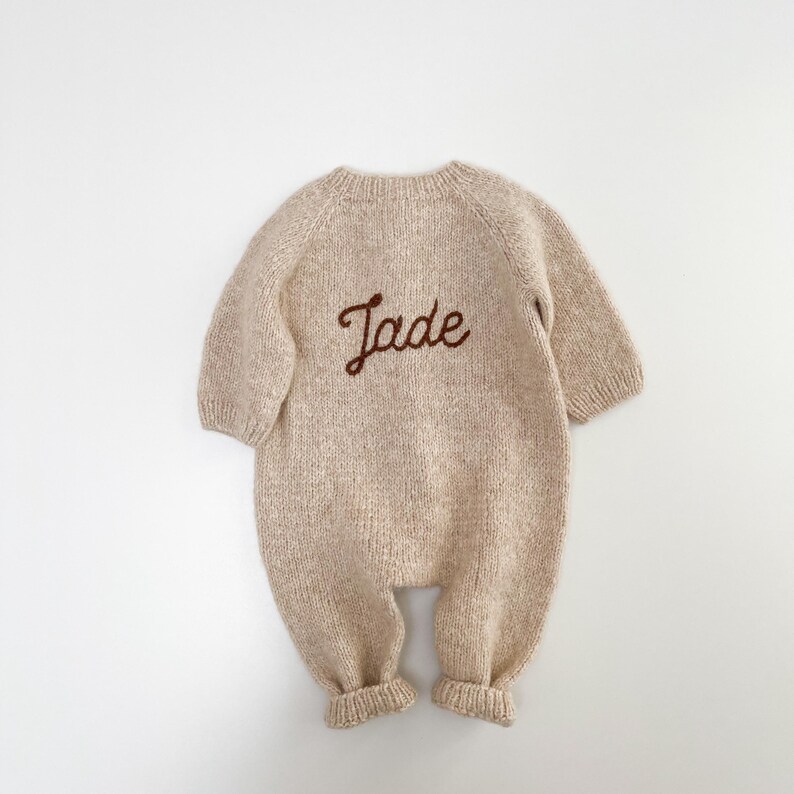 May include: A beige long-sleeved knitted romper featuring a round neckline and ribbed cuffs at the wrists and ankles. The center of the chest displays the name Jade embroidered in a dark brown cursive script, creating a clear contrast.