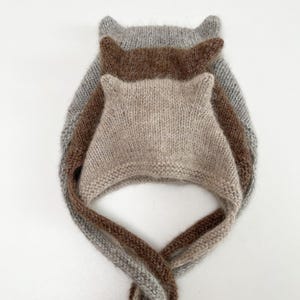 Könnte beinhalten: Ein Stapel aus drei gestrickten Hauben mit kleinen spitzen Ohren an der Oberseite. Die Hauben bestehen aus flauschigem Garn mit weicher Textur in den Farben Hellgrau, Mittelbraun und Beige. Jede Haube hat lange, gestrickte Bindebänder.