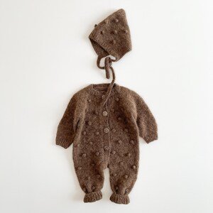 Könnte beinhalten: Ein braunes Strick-Baby-Set, bestehend aus einem Strampler und einer passenden Mütze. Der Strampler hat eine Knopfleiste und ist mit kleinen, strukturierten Bommeln verziert. Die Mütze hat Bindebänder und ähnliche Bommeln. Das Set ist aus weichem, strukturiertem Material.