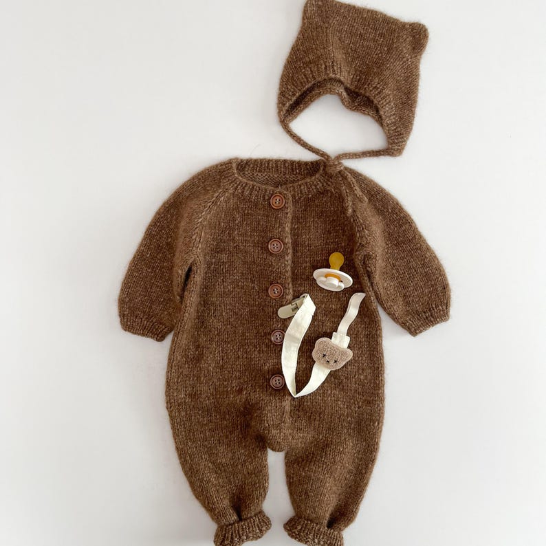 May include: A brown knitted long-sleeve romper with a front button closure, featuring a matching bonnet with small ear-like protrusions. A white pacifier clip with a small brown bear-shaped detail and a white pacifier are placed on the front.