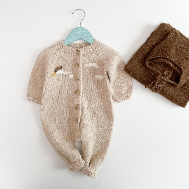 May include: A beige knitted long-sleeve romper with a button-down front, featuring a small embroidered cloud design on the chest. Beside it lies a matching dark brown knitted bonnet with two visible buttons on the front strap.