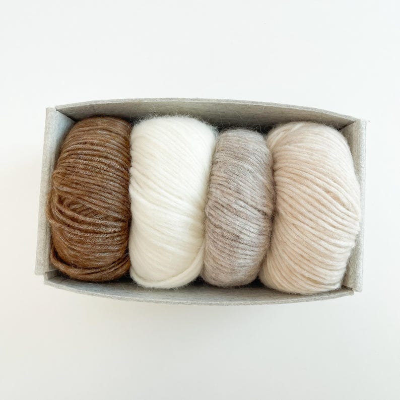 May include: Four skeins of yarn arranged in a rectangular felt container. The yarn colors include a dark brown, a bright white, a light taupe, and a soft cream. Each skein is wound into a rounded shape, filling the grey felt box completely.