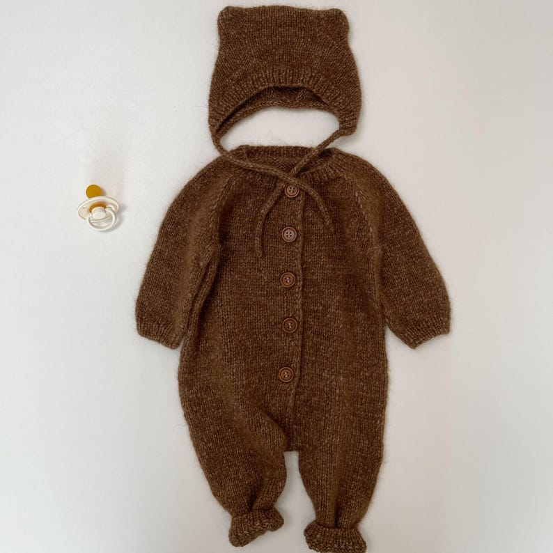 May include: A brown knitted long-sleeve romper with five matching buttons down the front center and a coordinating knitted bonnet with ties. A small white and yellow pacifier is positioned to the left of the knitted garment set.