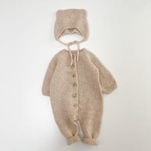May include: A beige knitted long-sleeve romper with a front button closure and a matching bonnet featuring small rounded ears on top. The garment is crafted from a textured yarn, showcasing a consistent neutral color and ribbed cuffs at the wrists.