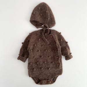 Peut inclure: Une barboteuse à manches longues en tricot marron foncé avec un motif texturé à pompons sur tout le tissu. Le vêtement comprend une fermeture latérale à cinq petits boutons marron, des poignets côtelés et un bonnet pointu assorti à nouer.