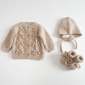 Peut inclure: Ensemble de bébé tricoté beige, comprenant un cardigan boutonné avec des détails de boules, un bonnet avec des liens à nouer et des chaussons assortis. L'ensemble est fait d'un fil doux et texturé. La couleur neutre convient à tous les bébés.