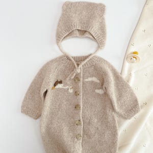 May include: A beige knit long-sleeve romper with a button-down front and a matching bonnet with ear-like details. The romper features embroidered white cloud motifs and a small stork carrying a bundle. A cream-colored pacifier is placed nearby.