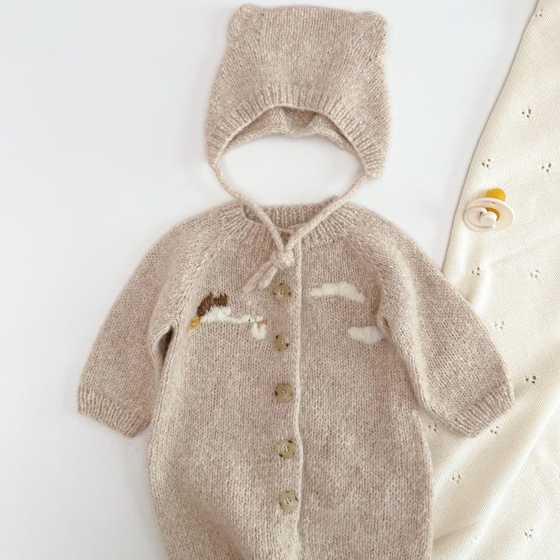 May include: A beige knit long-sleeve romper with a button-down front and a matching bonnet with ear-like details. The romper features embroidered white cloud motifs and a small stork carrying a bundle. A cream-colored pacifier is placed nearby.