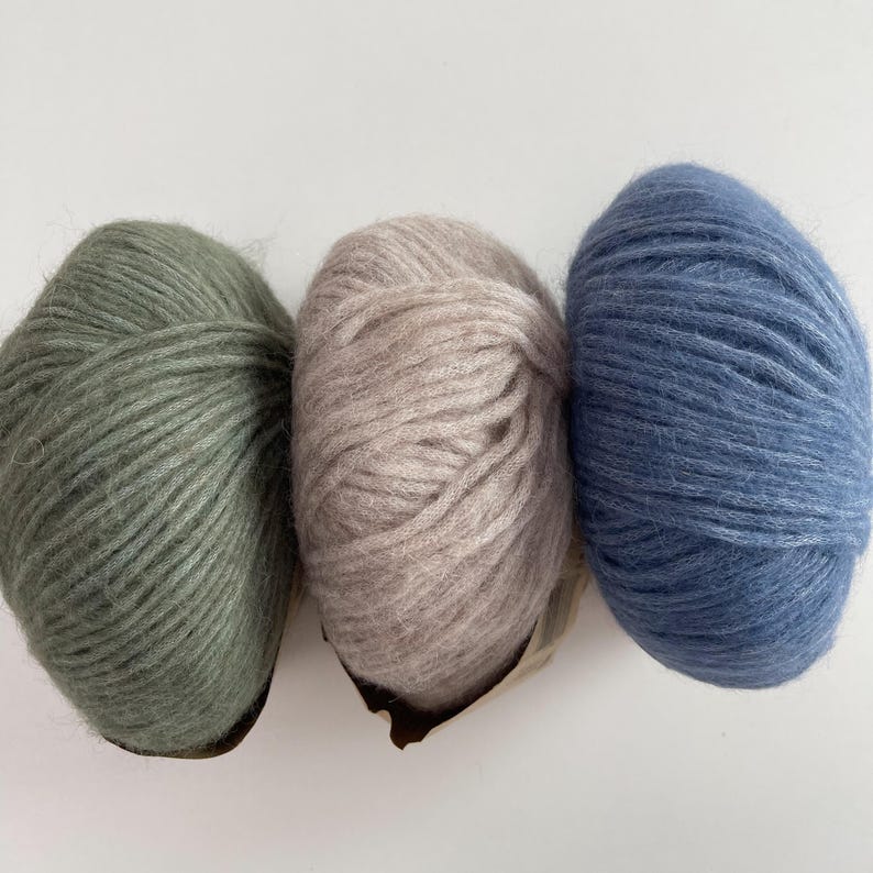 May include: Three round balls of fuzzy yarn arranged in a horizontal row. The left ball is sage green, the center ball is a light beige or taupe, and the right ball is a medium blue. Each ball features a soft, textured, and slightly hairy fiber.