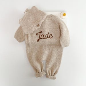 May include: A beige knitted long-sleeve romper and matching bonnet set. The romper features the name Jade embroidered in dark brown cursive script across the chest. The garment has ribbed cuffs at the wrists and ankles, with a simple round neckline.