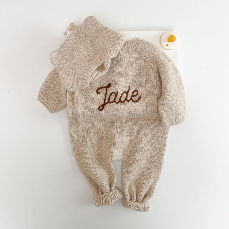 May include: A beige knitted long-sleeve romper and matching bonnet set. The romper features the name Jade embroidered in dark brown cursive script across the chest. The garment has ribbed cuffs at the wrists and ankles, with a simple round neckline.