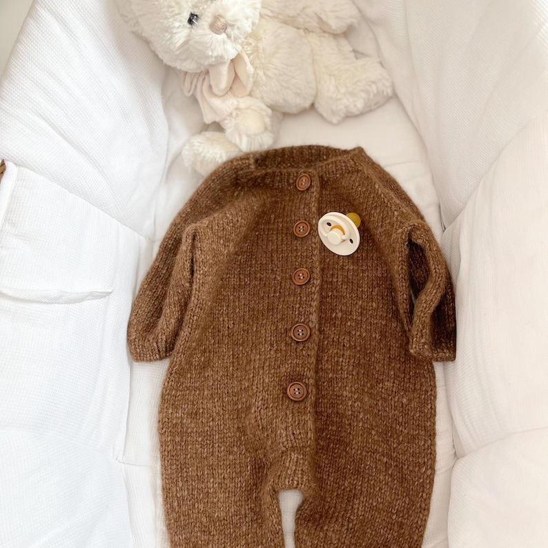 May include: A brown long-sleeved knitted romper featuring a round neckline and five brown buttons down the front. A cream-colored pacifier is attached to the chest area of the garment, and a white plush teddy bear is positioned above the romper.