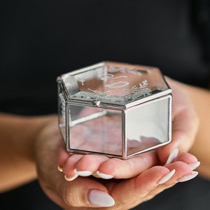 May include: A clear glass ring box with a silver metal frame. The box has a hexagonal shape and is decorated with a floral design.