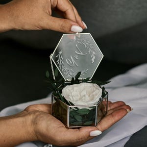 May include: A hexagonal glass ring box with a silver frame, containing a white rose and greenery. The lid is engraved with initials and a floral design. The box is held open by a person's hand.