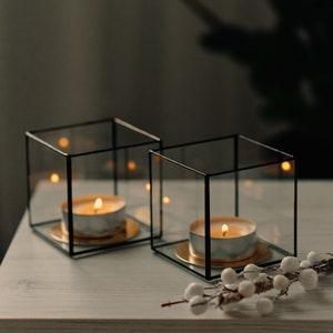 Può includere: Due portacandele in vetro con cornici nere e basi dorate, ciascuna con una candela a tè accesa. I portacandele sono su una superficie di legno bianco con un ramo bianco con piccole bacche bianche.