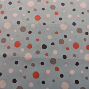 May include: Close-up of a light blue fabric with a scattered polka dot pattern. The dots vary in size and color, including orange, white, gray, and black. The fabric appears to be cotton or a similar material.
