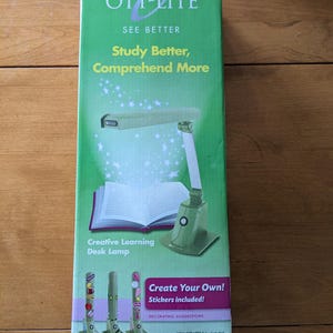 May include: A green and white desk lamp with a flexible arm and a magnifying glass. The lamp is on a white book with the text "Creative Learning Desk Lamp". The box also includes the text "OTT-LITE See Better Study Better, Comprehend More" and "Create Your Own! Stickers included!"