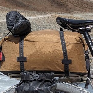 Puede incluir: Una bolsa de bicicleta marr&oacute;n impermeable con correas negras est&aacute; unida a un portabultos negro en una bicicleta. La bicicleta tiene un sill&iacute;n negro y un manillar negro con un logotipo Olsen negro y blanco.