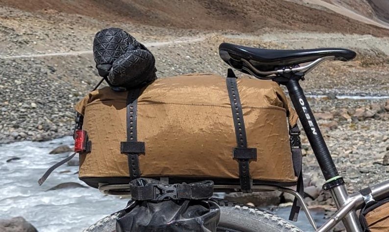 Puede incluir: Una bolsa de bicicleta marr&oacute;n impermeable con correas negras est&aacute; unida a un portabultos negro en una bicicleta. La bicicleta tiene un sill&iacute;n negro y un manillar negro con un logotipo Olsen negro y blanco.