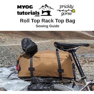 Puede incluir: Una bolsa de equipaje de bicicleta marr&oacute;n con cierre de rollo en la parte superior, fijada a una bicicleta. La bolsa tiene m&uacute;ltiples correas y hebillas. La bicicleta est&aacute; aparcada en un camino de tierra cerca de un r&iacute;o.