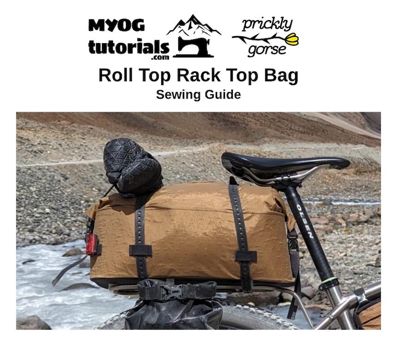 Puede incluir: Una bolsa de equipaje de bicicleta marr&oacute;n con cierre de rollo en la parte superior, fijada a una bicicleta. La bolsa tiene m&uacute;ltiples correas y hebillas. La bicicleta est&aacute; aparcada en un camino de tierra cerca de un r&iacute;o.