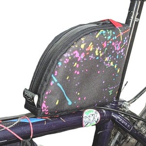 May include: A black bike frame with a colorful, splatter-painted handlebar bag attached. The bag has a zipper closure and a black strap.