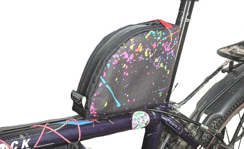 May include: A black bike frame with a colorful, splatter-painted handlebar bag attached. The bag has a zipper closure and a black strap.