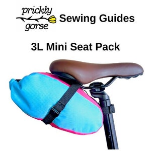 May include: A blue and pink bike seat pack attached to a brown bike seat. The pack is labelled "3L Mini Seat Pack".