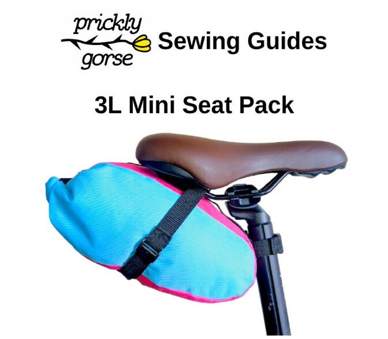 Może przedstawiać: Niebiesko-r&oacute;żowa torba Mini Seat Pack 3L przymocowana do siodełka rowerowego. Torba jest zabezpieczona czarnym paskiem. Tekst "prickly gorse Sewing Guides" i "3L Mini Seat Pack" są widoczne.