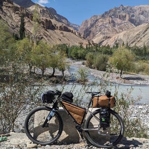 Puede incluir: Una bicicleta de touring plateada con alforjas marrones y una bolsa de manillar negra est&aacute; aparcada en un camino de tierra cerca de un r&iacute;o en un paisaje monta&ntilde;oso.
