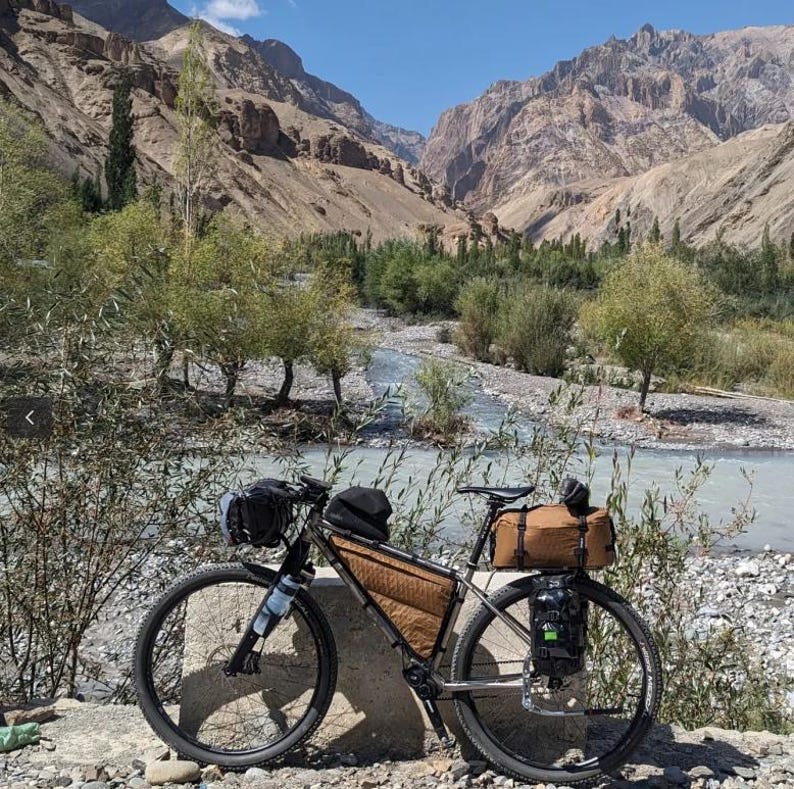 Puede incluir: Una bicicleta de touring plateada con alforjas marrones y una bolsa de manillar negra est&aacute; aparcada en un camino de tierra cerca de un r&iacute;o en un paisaje monta&ntilde;oso.