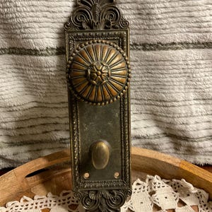 May include: An antique brass door plate and knob set featuring an ornate, rectangular backplate with intricate scrollwork at the top and bottom. The round, fluted door knob is centered above a smaller, oval-shaped thumb turn mechanism.