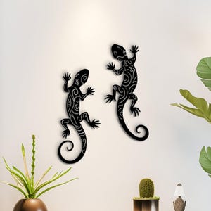 May include: Two black metal lizard wall art pieces with detailed tribal patterns are displayed on a white wall. The lizards feature intricate designs and curled tails. The scene includes a console table with plants and decorative items, creating a modern interior design.